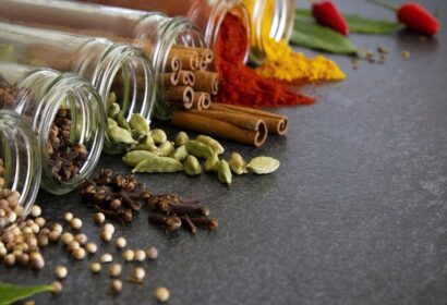 Various spices, including cinnamon sticks, cardamom pods, cloves, bay leaves, and chili peppers, spilling from glass jars onto a gray surface.