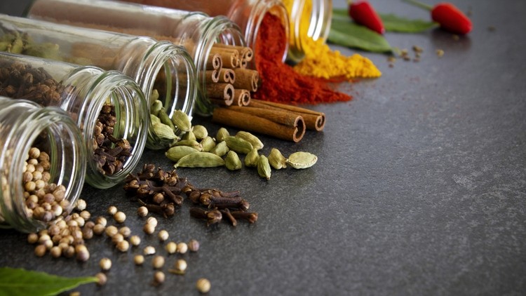 Various spices, including cinnamon sticks, cardamom pods, cloves, bay leaves, and chili peppers, spilling from glass jars onto a gray surface.