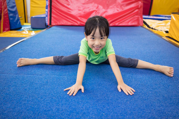 A child in a green shirt and gray leggings is doing a full split on a blue mat in a colorful gym or play area.