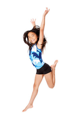 A girl in a blue and white leotard and black shorts jumps with arms raised and one leg bent, smiling against a white background.