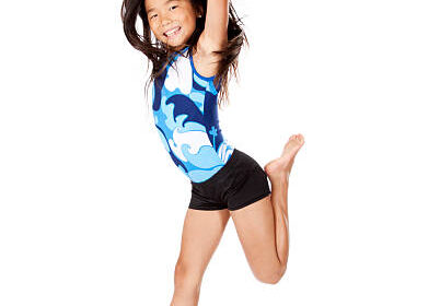 A girl in a blue and white leotard and black shorts jumps with arms raised and one leg bent, smiling against a white background.