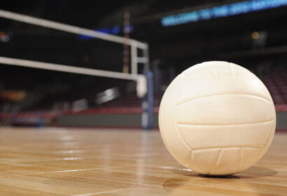 Close-up of a volleyball on a polished wooden court with a net and blurred arena seats in the background.