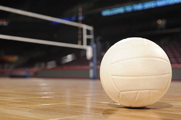 Close-up of a volleyball on a polished wooden court with a net and blurred arena seats in the background.