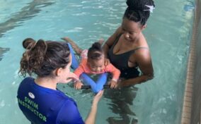 A swim instructor and a woman hold a baby using a kickboard in a swimming pool.