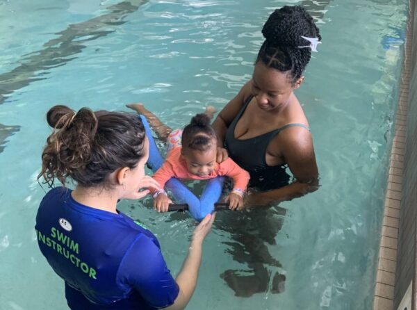 A swim instructor and a woman hold a baby using a kickboard in a swimming pool.