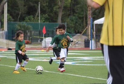 Two young children playing soccer on a field, wearing green jerseys and black shorts, with one adult in a yellow jersey nearby.