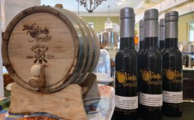 Small wooden barrel with "Olive Amelia" branding next to three bottles of balsamic vinegar on a countertop in a store.