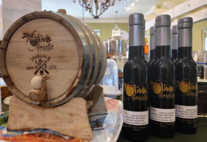 Small wooden barrel with "Olive Amelia" branding next to three bottles of balsamic vinegar on a countertop in a store.