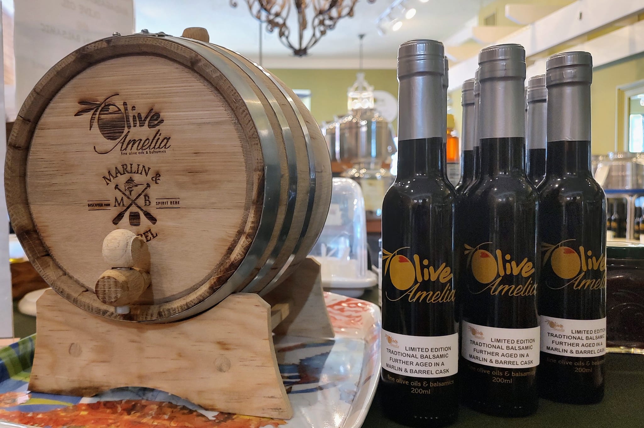 Small wooden barrel with "Olive Amelia" branding next to three bottles of balsamic vinegar on a countertop in a store.