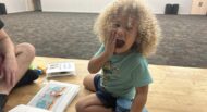 A young child with curly hair sits on the floor, playfully covering their mouth with one hand, surrounded by books and toys, in a large, empty room.