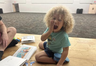 A young child with curly hair sits on the floor, playfully covering their mouth with one hand, surrounded by books and toys, in a large, empty room.