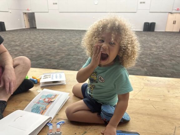 A young child with curly hair sits on the floor, playfully covering their mouth with one hand, surrounded by books and toys, in a large, empty room.