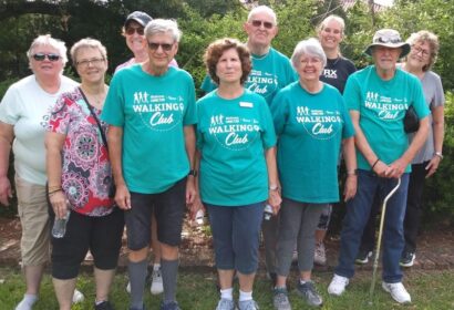 A group of twelve people standing outdoors in a park. Most are wearing matching green "Walking Club" T-shirts.