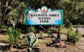 A green and white sign at the entrance of Kathryn Abbey Hanna Park surrounded by plants and trees on a sunny day.