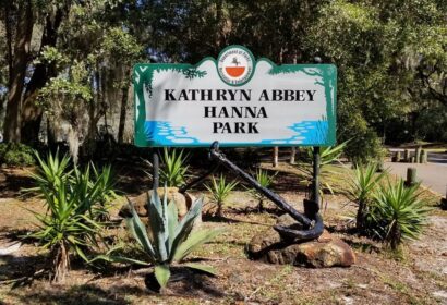 A green and white sign at the entrance of Kathryn Abbey Hanna Park surrounded by plants and trees on a sunny day.