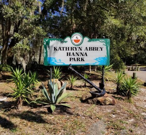 A green and white sign at the entrance of Kathryn Abbey Hanna Park surrounded by plants and trees on a sunny day.