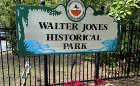 Sign reading "Walter Jones Historical Park" in front of a black fence and trees, with pink flowers at the base. The sign includes a logo for the Department of Parks, Recreation & Entertainment.