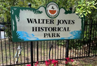 Sign reading "Walter Jones Historical Park" in front of a black fence and trees, with pink flowers at the base. The sign includes a logo for the Department of Parks, Recreation & Entertainment.