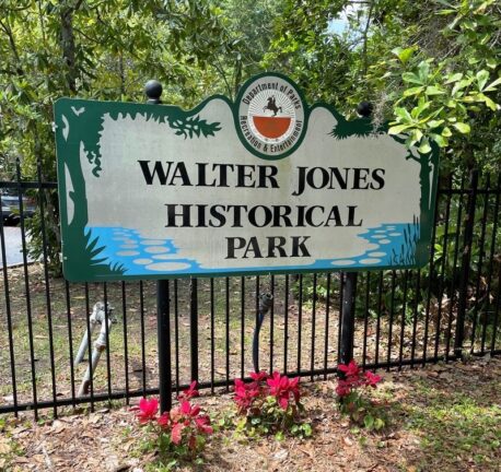 Sign reading "Walter Jones Historical Park" in front of a black fence and trees, with pink flowers at the base. The sign includes a logo for the Department of Parks, Recreation & Entertainment.