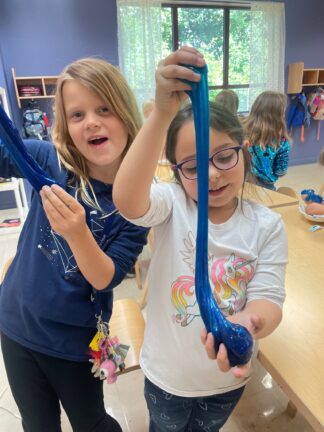 Two girls in a classroom stretch blue slime with their hands. One girl wears glasses and a unicorn shirt, while the other has her mouth open and is wearing a dark blue shirt. Other children are in the background.