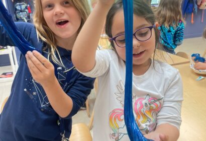 Two girls in a classroom stretch blue slime with their hands. One girl wears glasses and a unicorn shirt, while the other has her mouth open and is wearing a dark blue shirt. Other children are in the background.