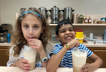 Two children are drinking beverages through straws at a counter. The girl on the left has curly hair and wears a blue headband, while the boy on the right is smiling and wearing a striped shirt.