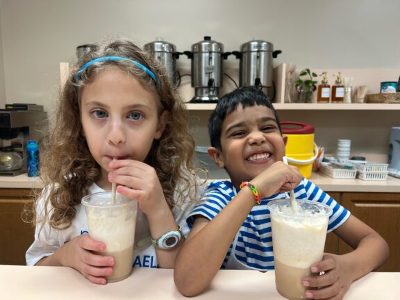 Two children are drinking beverages through straws at a counter. The girl on the left has curly hair and wears a blue headband, while the boy on the right is smiling and wearing a striped shirt.