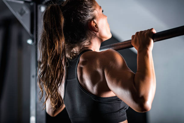 A woman with a ponytail performs a pull-up on a bar at a gym, wearing a black sports bra.