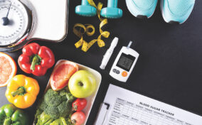 A table with a weighing scale, vegetables, fruits, dumbbells, measuring tape, sneakers, glucose meter displaying 5.6, and a clipboard with a blood sugar tracker.