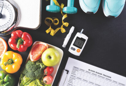 A table with a weighing scale, vegetables, fruits, dumbbells, measuring tape, sneakers, glucose meter displaying 5.6, and a clipboard with a blood sugar tracker.