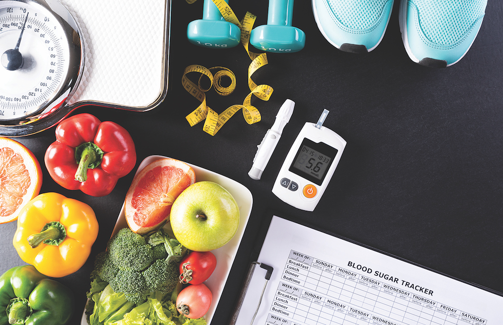 A table with a weighing scale, vegetables, fruits, dumbbells, measuring tape, sneakers, glucose meter displaying 5.6, and a clipboard with a blood sugar tracker.