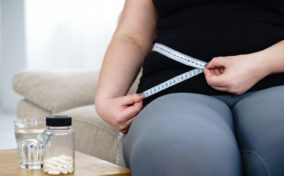 Person measuring their waist with a tape measure while seated. Nearby, there is a small table with a bottle of pills and a glass of water on it.