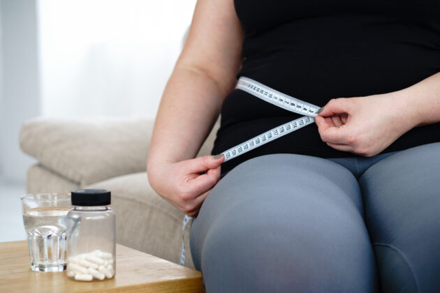 Person measuring their waist with a tape measure while seated. Nearby, there is a small table with a bottle of pills and a glass of water on it.