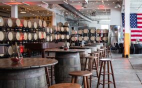 A modern brewery interior with wooden barrels stacked against the wall, high-top barrel tables with stools, string lights hanging from the ceiling, and an American flag displayed on the right.