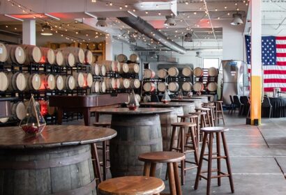 A modern brewery interior with wooden barrels stacked against the wall, high-top barrel tables with stools, string lights hanging from the ceiling, and an American flag displayed on the right.