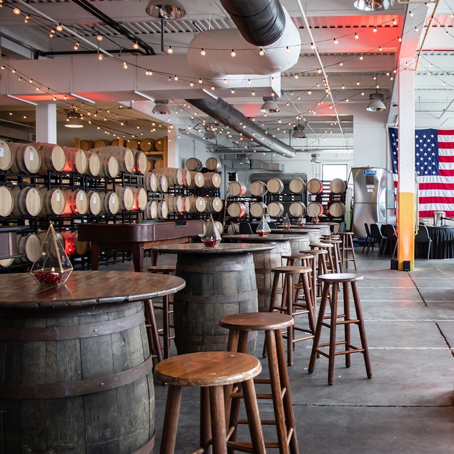 A modern brewery interior with wooden barrels stacked against the wall, high-top barrel tables with stools, string lights hanging from the ceiling, and an American flag displayed on the right.