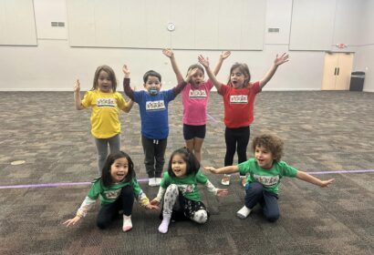 Eight children in colorful shirts enthusiastically pose with their arms raised in a large, empty room with a purple line on the floor.