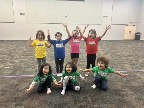 Eight children in colorful shirts enthusiastically pose with their arms raised in a large, empty room with a purple line on the floor.