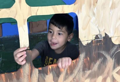 A young child looks out of a painted cutout window in a playhouse, holding a small object in their hand.