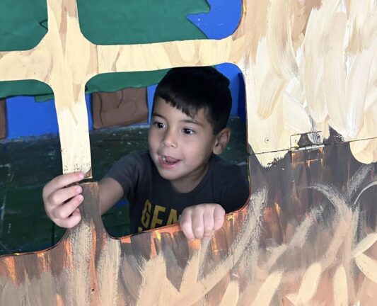 A young child looks out of a painted cutout window in a playhouse, holding a small object in their hand.