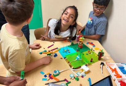 Children gather around a table building with colorful LEGO bricks, smiling and laughing. A laptop and LEGO box are also on the table.