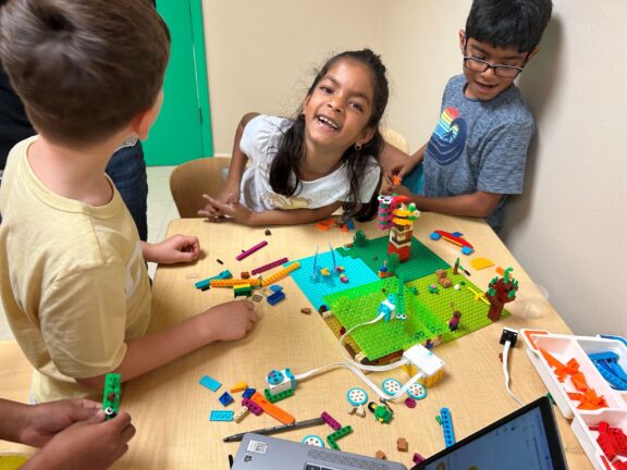 Children gather around a table building with colorful LEGO bricks, smiling and laughing. A laptop and LEGO box are also on the table.