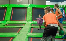 Children playing a game involving a ball on a trampoline court. The participants are wearing colorful shirts and are engaged in activities on the green and black trampolines.