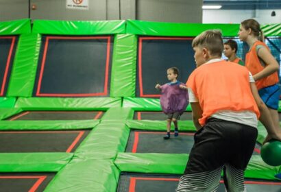 Children playing a game involving a ball on a trampoline court. The participants are wearing colorful shirts and are engaged in activities on the green and black trampolines.