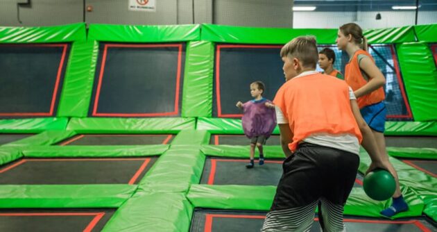 Children playing a game involving a ball on a trampoline court. The participants are wearing colorful shirts and are engaged in activities on the green and black trampolines.