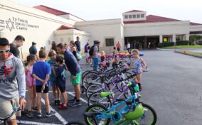 A group of people, including adults and children, gather outside a community center, with several bicycles lined up in the foreground. The building has a sign reading "Ed Parker Jewish Community Campus.