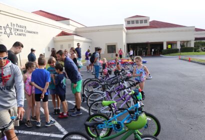 Children and adults gather outside a building by rows of parked bicycles, preparing for a community event. The building's sign says, "Ed Parker Jewish Community Campus.