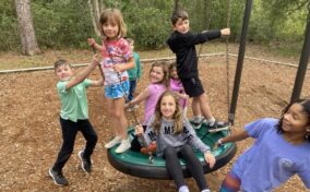 A group of six children, five girls and one boy, play on a saucer swing in an outdoor playground surrounded by trees. The children are smiling and appear to be enjoying themselves.