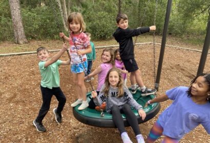 A group of six children, five girls and one boy, play on a saucer swing in an outdoor playground surrounded by trees. The children are smiling and appear to be enjoying themselves.