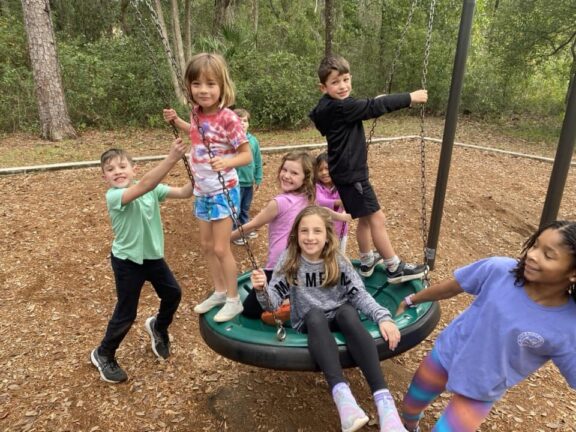 A group of six children, five girls and one boy, play on a saucer swing in an outdoor playground surrounded by trees. The children are smiling and appear to be enjoying themselves.